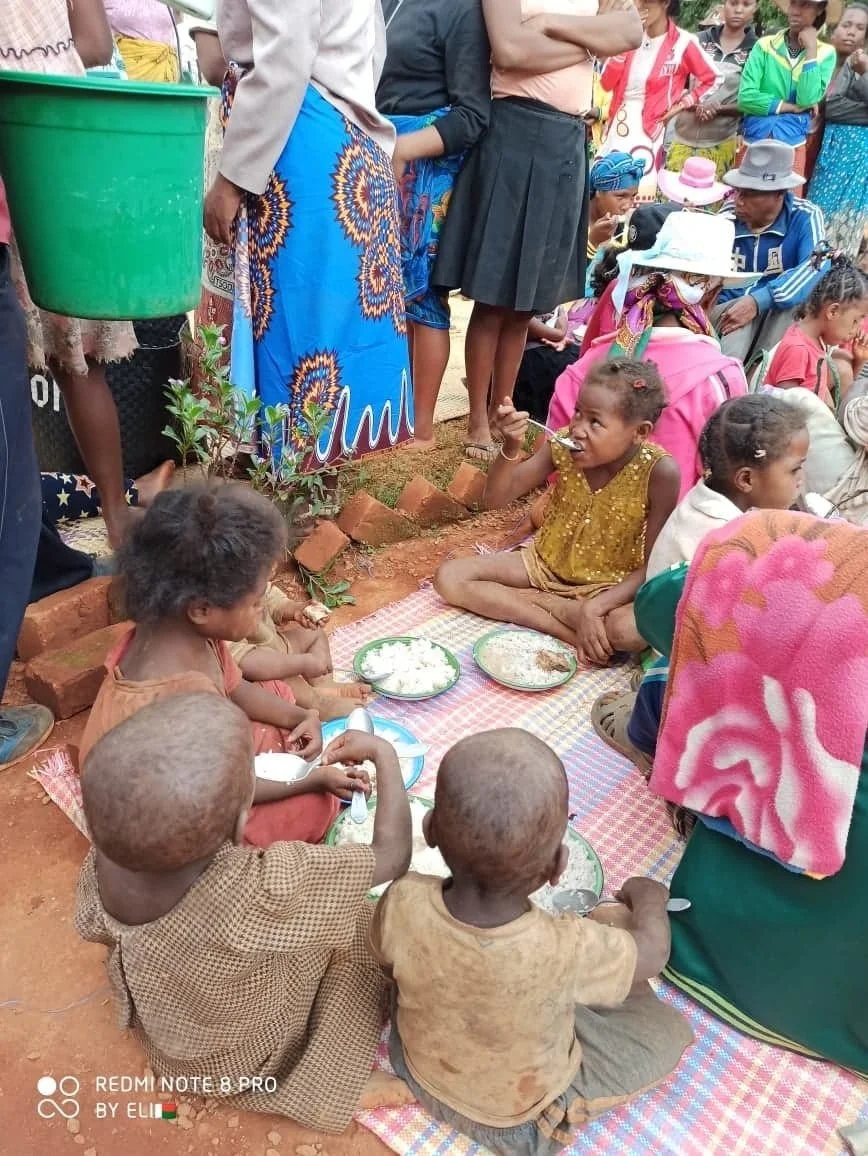 Children eating food on a mat outdoors in Madagascar surrounded by adults and other children.