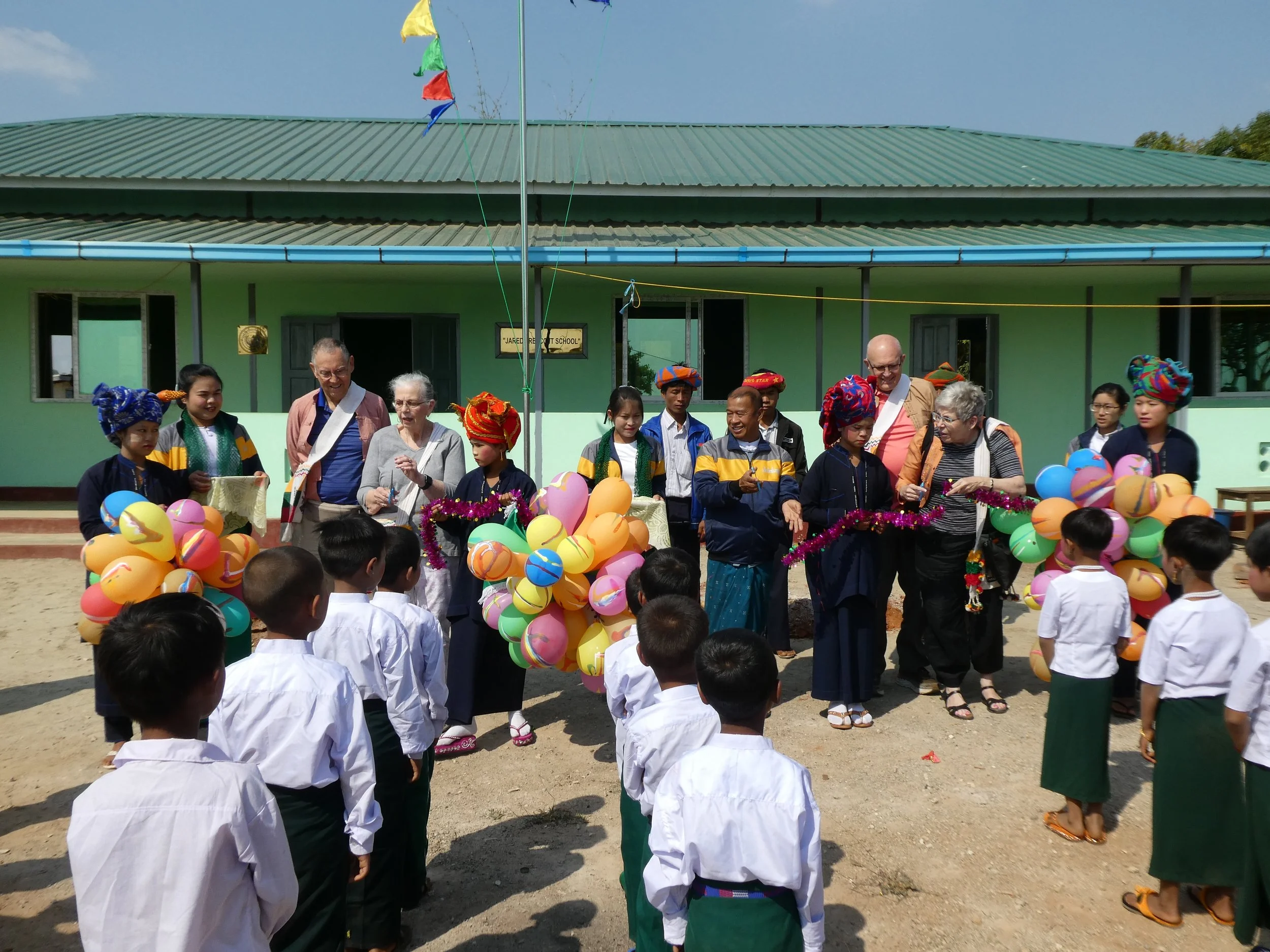 Group of children and adults standing outside a new  school building in Myanmar during a dedication celebration, with colorful balloons and decorations, some wearing traditional attire.