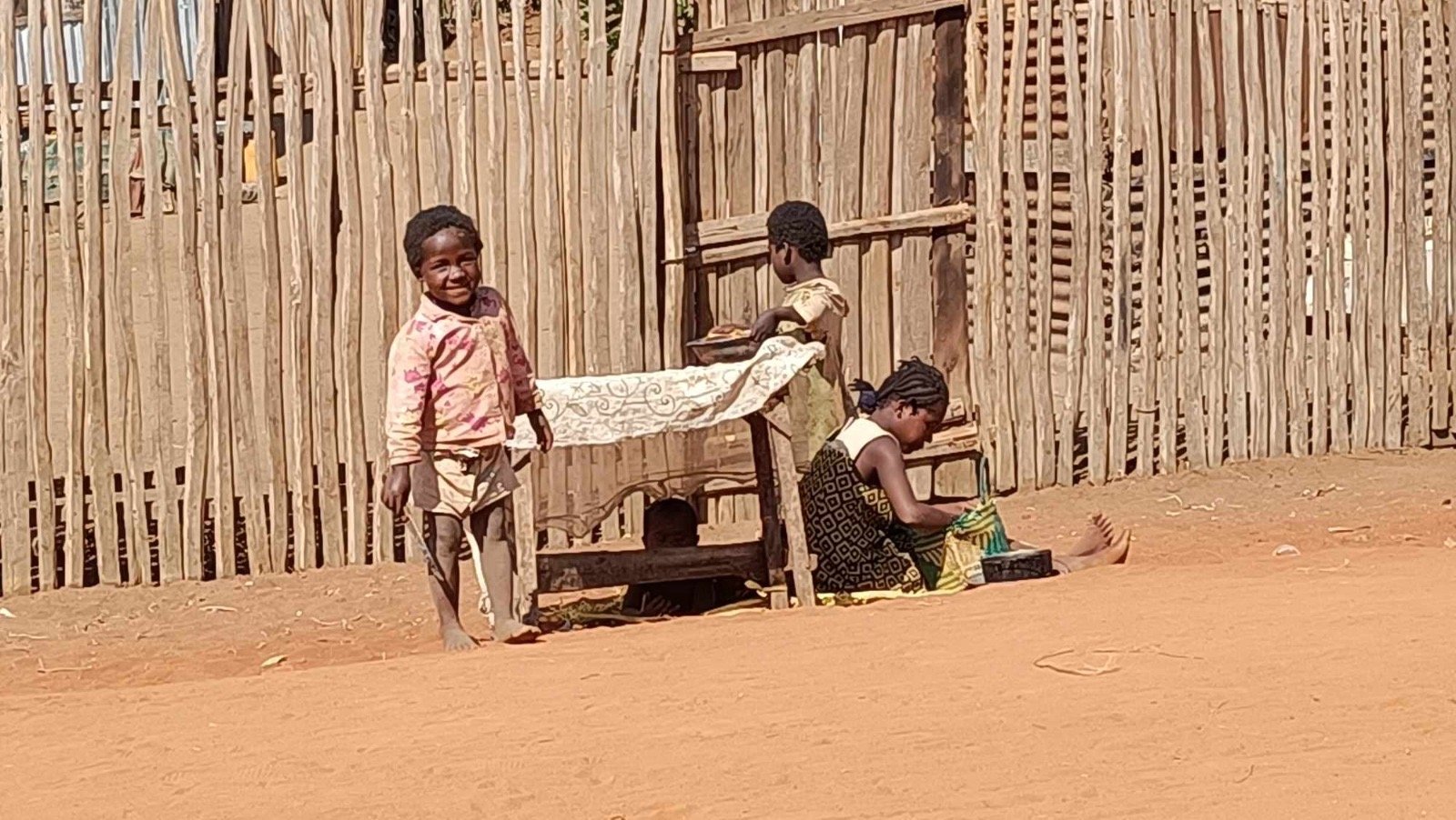Malagasy children playing and sitting in front of a wooden fence, with one girl smiling at the camera, two other children engaged in activities, and a sandy ground.