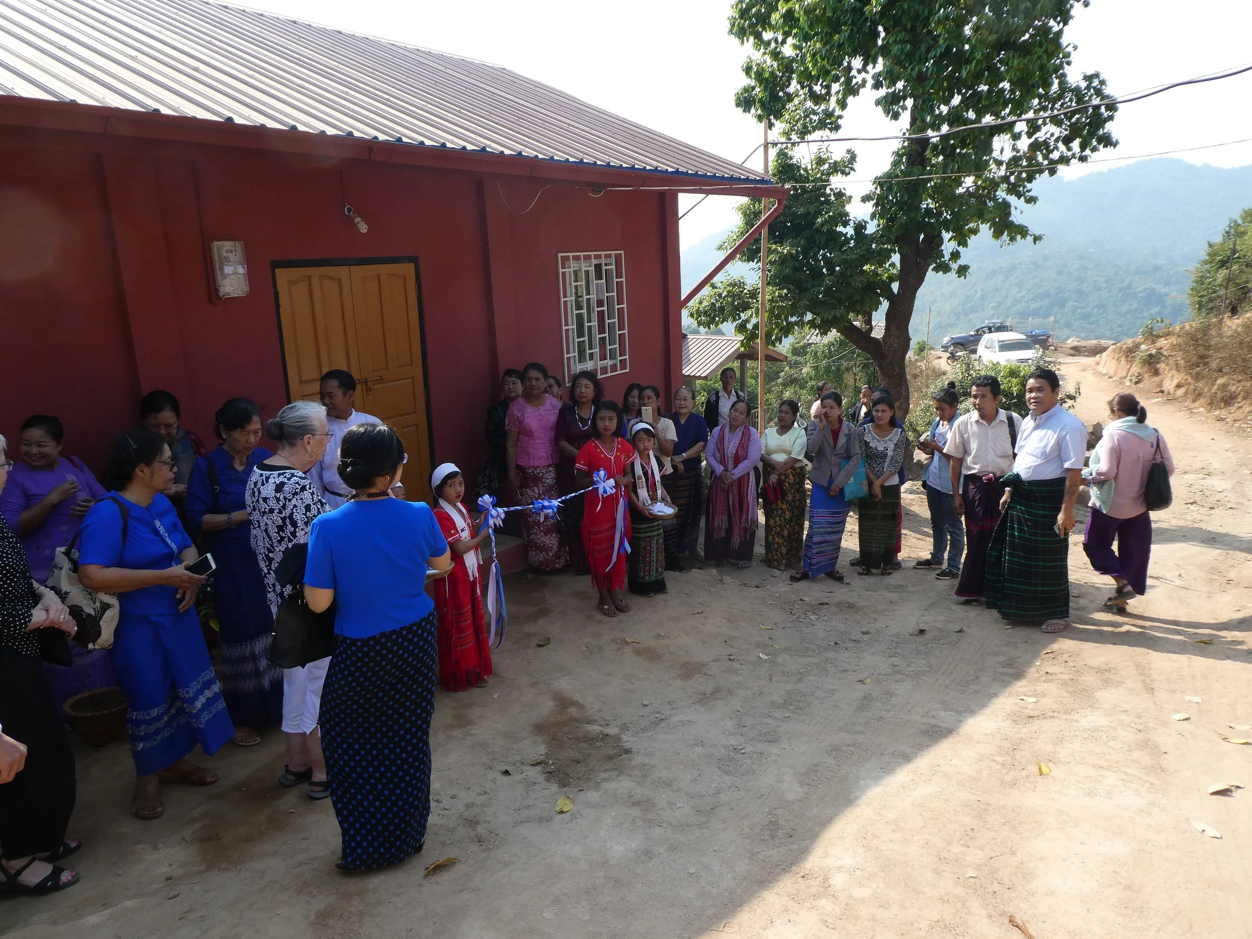 People gathered in front of a newly constructed  YWCA women's education centerin rural Myanmar, some wearing traditional clothing, during a ribbon-cutting ceremony outdoors.