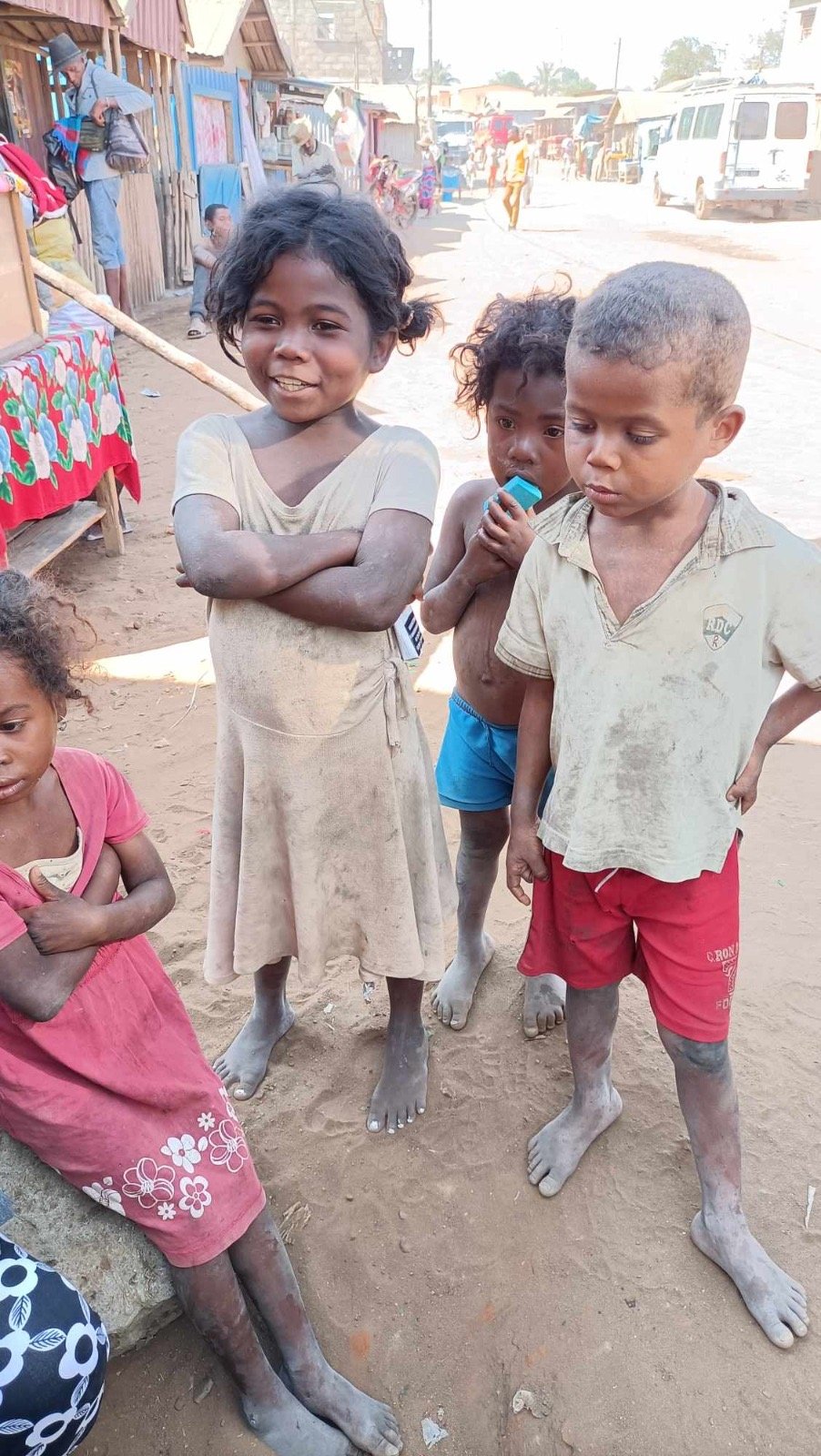 Children standing on dirt street in an impoverished area in Madagascar, some with dust on their skin and clothes, with makeshift shops and people in the background.