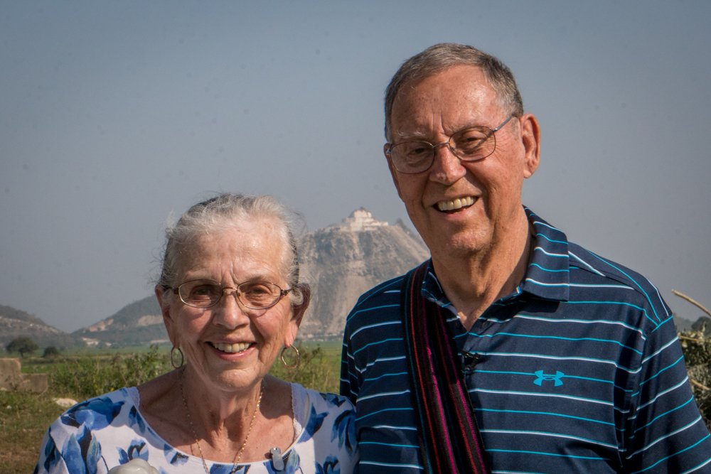 An elderly woman and man smiling outdoors in Myanmar with a mountain in the background.