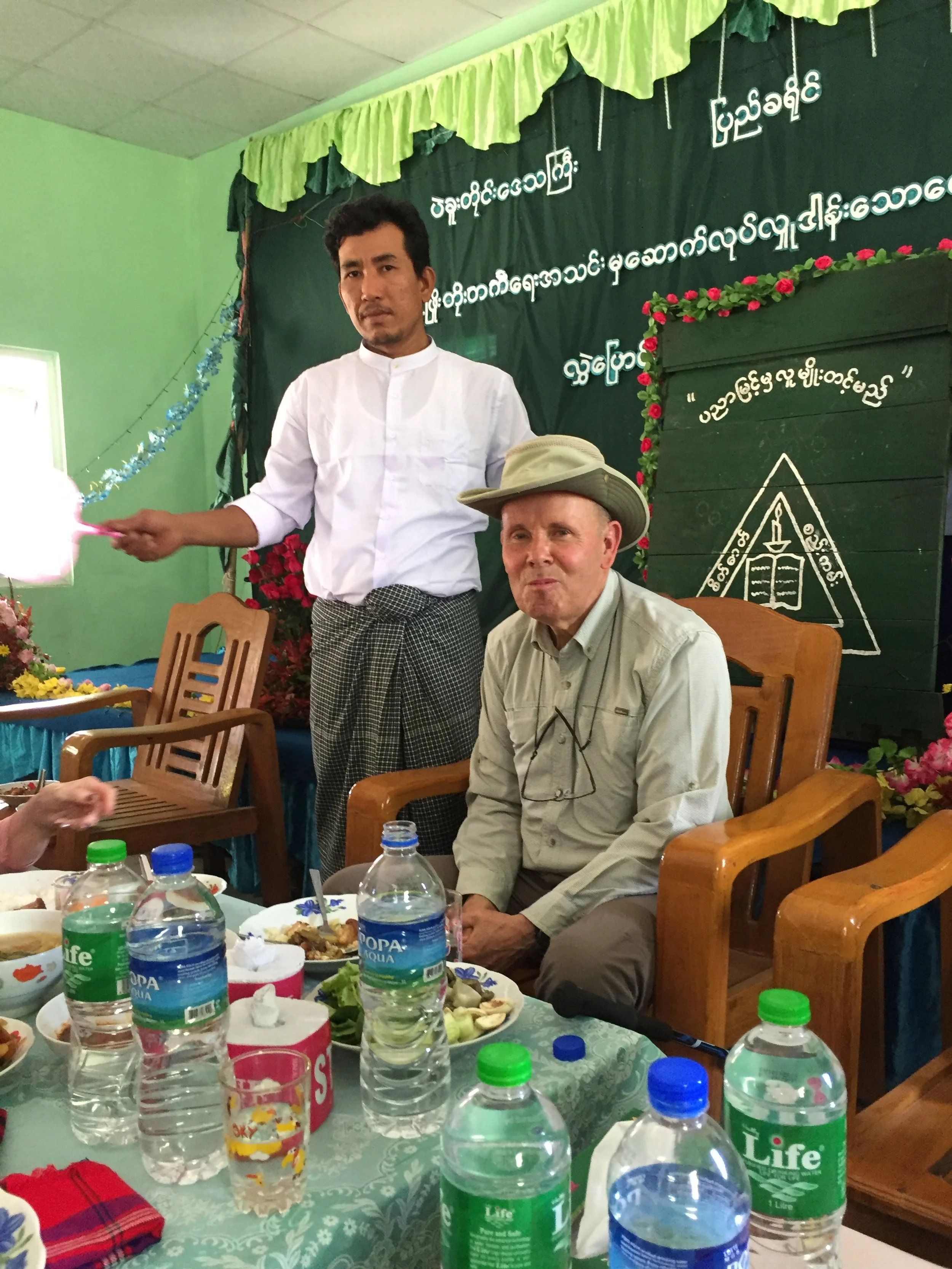 Two men are seated and standing at a table during a celebration, with a green backdrop with white Burmese script and floral decorations behind them. One man is wearing a light-colored shirt and a hat, and the other man is standing behind him wearing a white shirt and patterned sarong, holding a lit candle or wand. There are water bottles and food on the table in front of them.