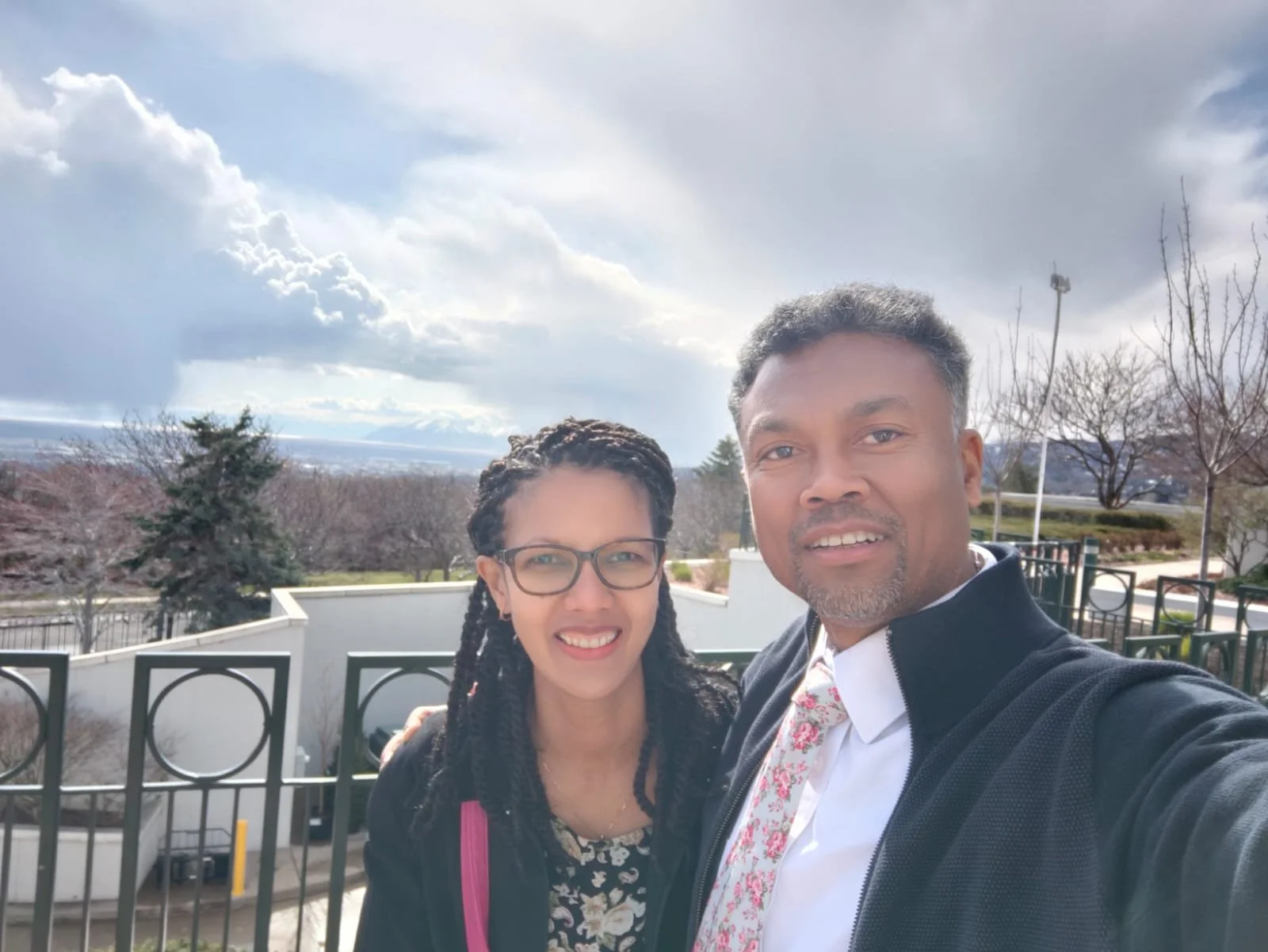 A man and woman taking a selfie outdoors during daytime, with trees, a fence, and cloudy sky in the background.