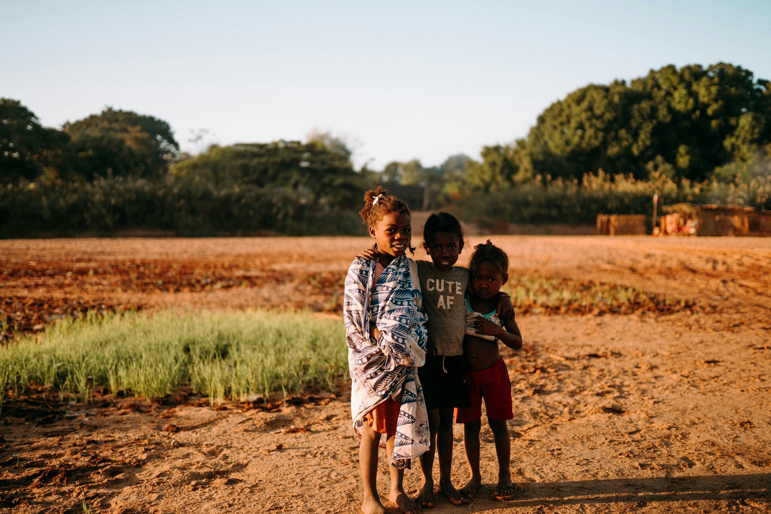 Three children standing together outdoors in a rural Madagawscar area during sunset, with a field and trees in the background.