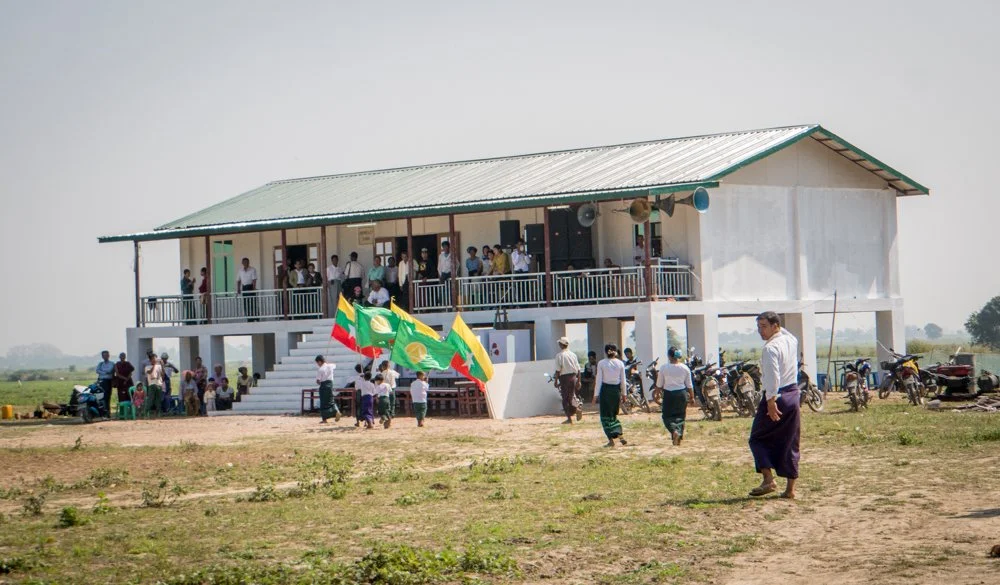 A gathering of people outside a new school provided by the Jared Prescott Trust , for its dedication, with some holding flags of Myanmar, in a rural area with motorcycles parked nearby.