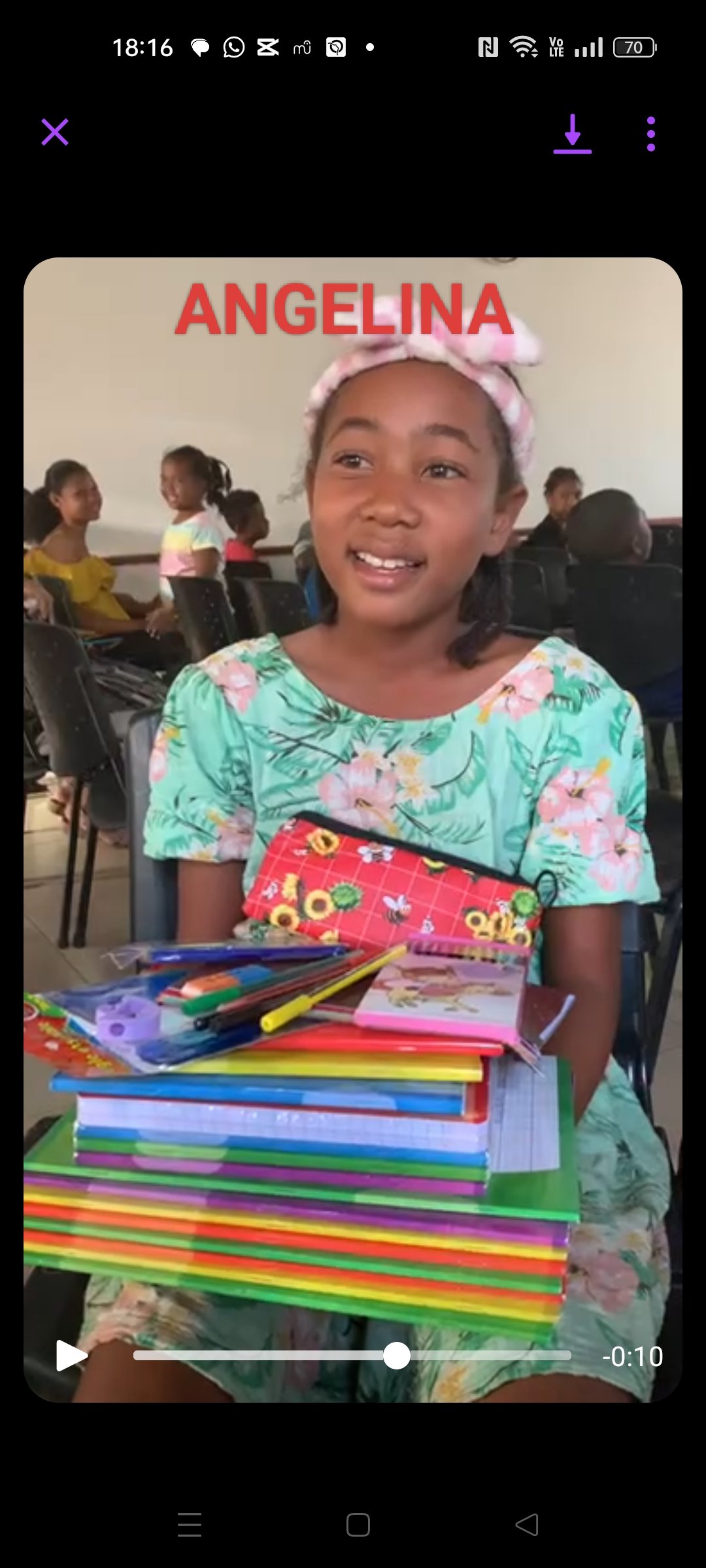 A young girl with a pink headband and floral dress sitting in a classroom, smiling, with a stack of colorful notebooks and stationery on her lap and chest. There are other children sitting in the background.