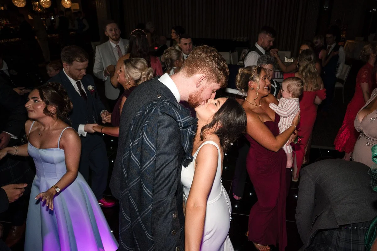 A group of people dancing and socialising at a wedding reception, with the bride and groom in the foreground sharing a kiss at Radstone Hotel