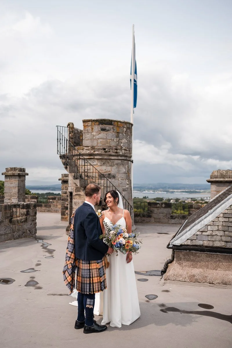 A bride and groom standing on Dundas Castle rooftop, holding hands and smiling at each other, with a Scottish flag flying in the background.