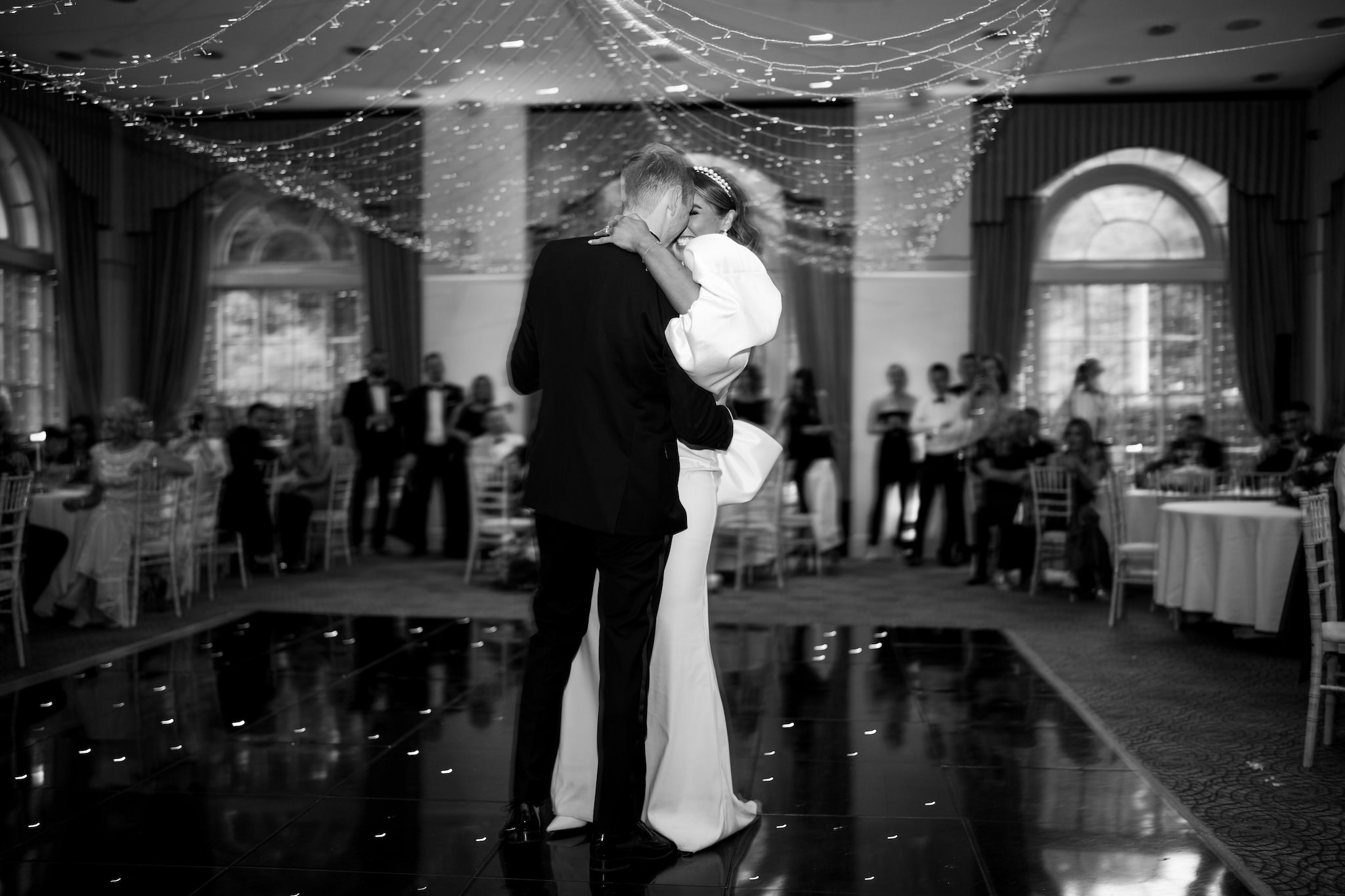 Black and white photo of a bride and groom dancing closely at a wedding reception at Balbirnie House, surrounded by seated guests and decorated with twinkling string lights hanging from the ceiling.