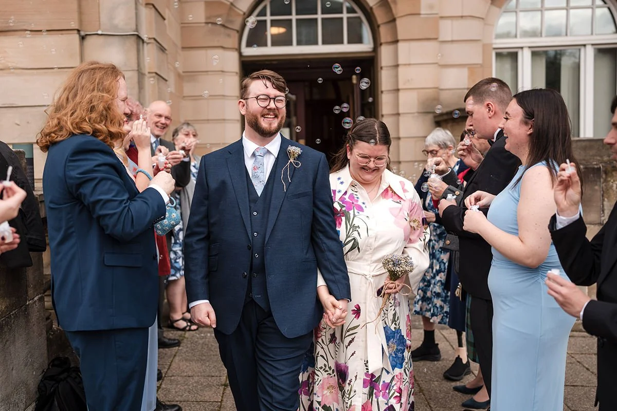 A joyful wedding celebration outside a Hamilton Registry Office, with a bride and groom holding hands and smiling, surrounded by friends and family blowing bubbles.