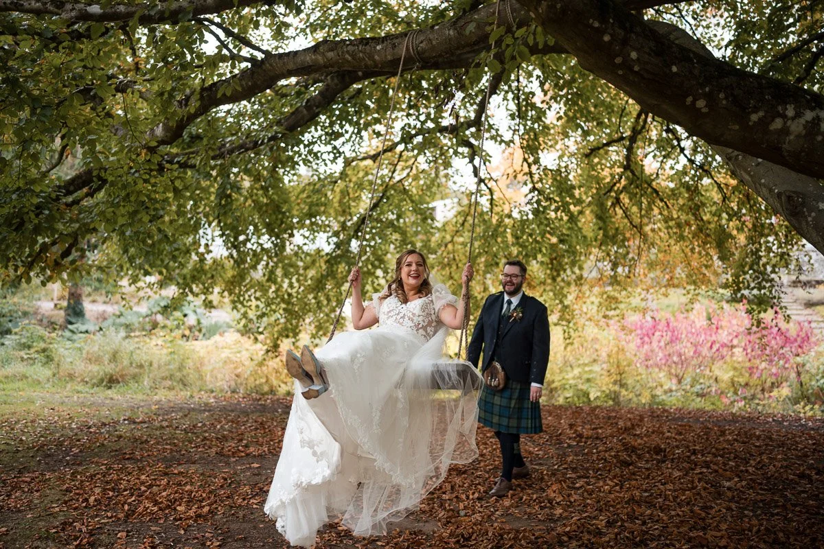 A bride in a white wedding dress joyfully swings on a tree swing, while a groom in a kilt watches and smiles in a park with autumn foliage at Rufflets, St. Andrews