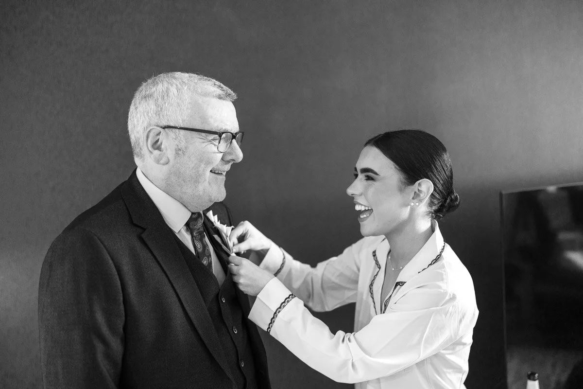 A bride helping a man put on a boutonniere in a formal setting, both smiling.