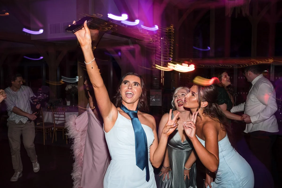 Group of young women taking a selfie and dancing at a wedding, with colourful lights and other guests in the background at Enterkine House Hotel