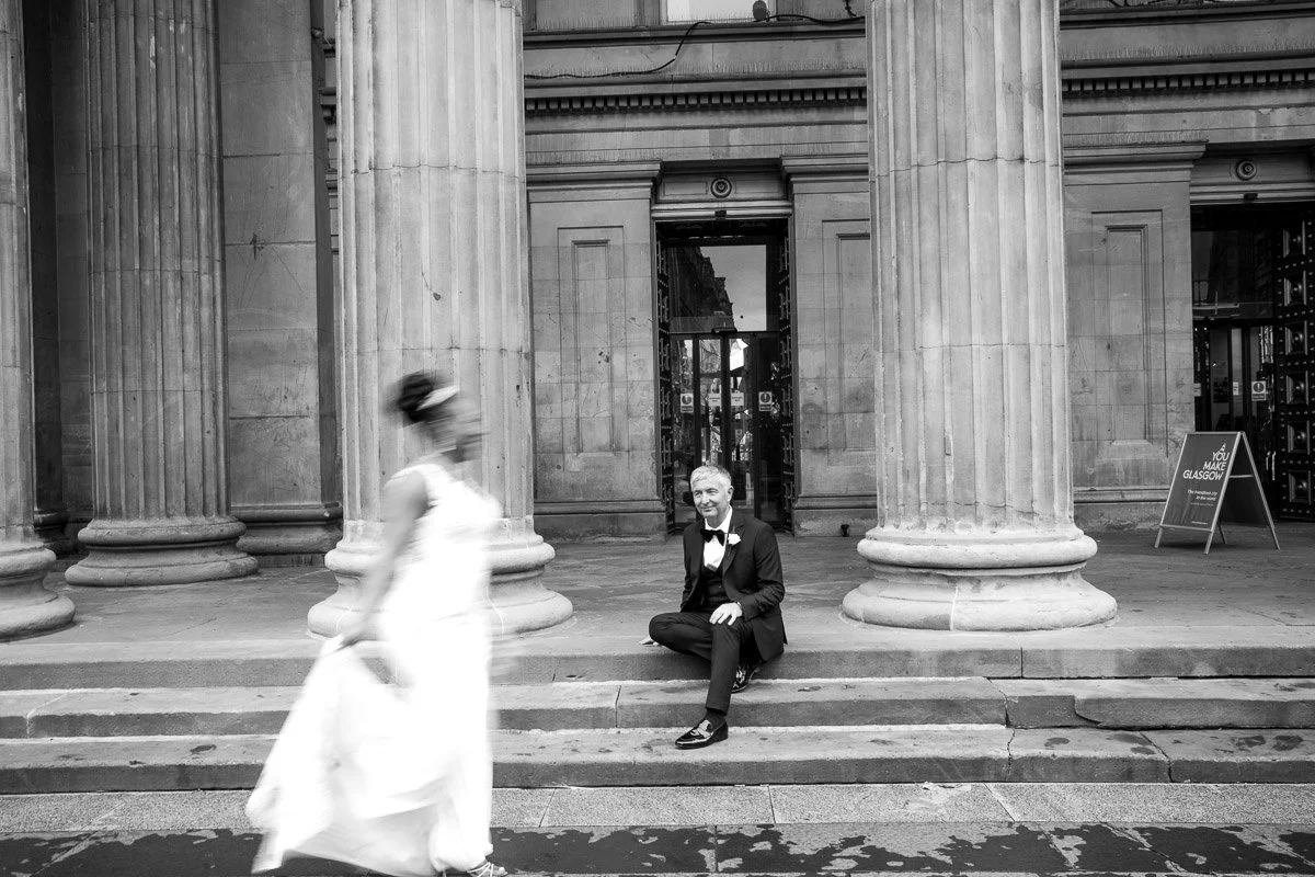 A broom in a tuxedo with a bow tie and a flower on his lapel is sitting on steps in front of large columns outside a building, while a bride in a white dress walks past in front of him after their Glasgow city centre elopement