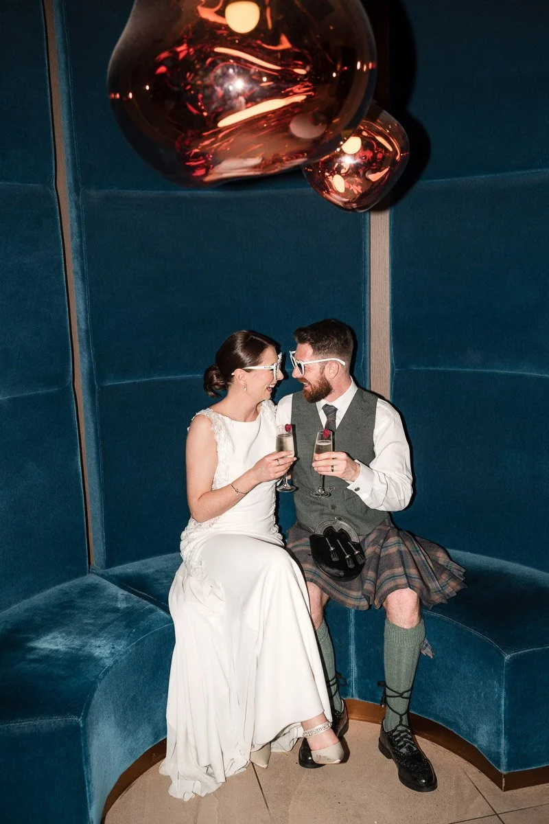 A bride and groom sitting in a booth, smiling and clinking champagne glasses. The bride is wearing a white wedding dress, and the groom is wearing a kilt with traditional Scottish attire. They are sharing a joyful moment at their wedding celebration.