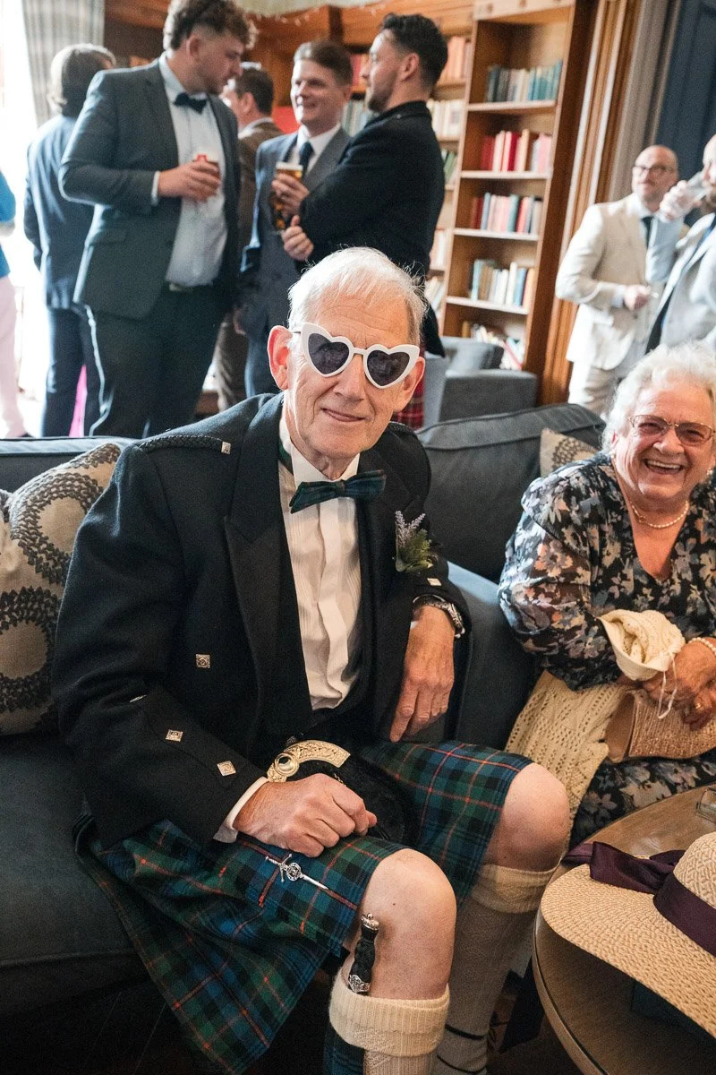 An elderly man wearing heart-shaped sunglasses, a tuxedo, a bow tie, and a kilt, sitting on a sofa at a wedding by smiling people at Balbirnie House