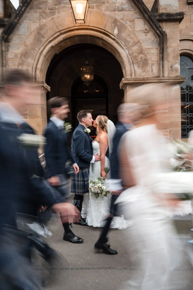 A bride and groom sharing a kiss outside a stone building with an arched doorway, surrounded by people walking past in motion blur at their wedding at Cornhill Castle