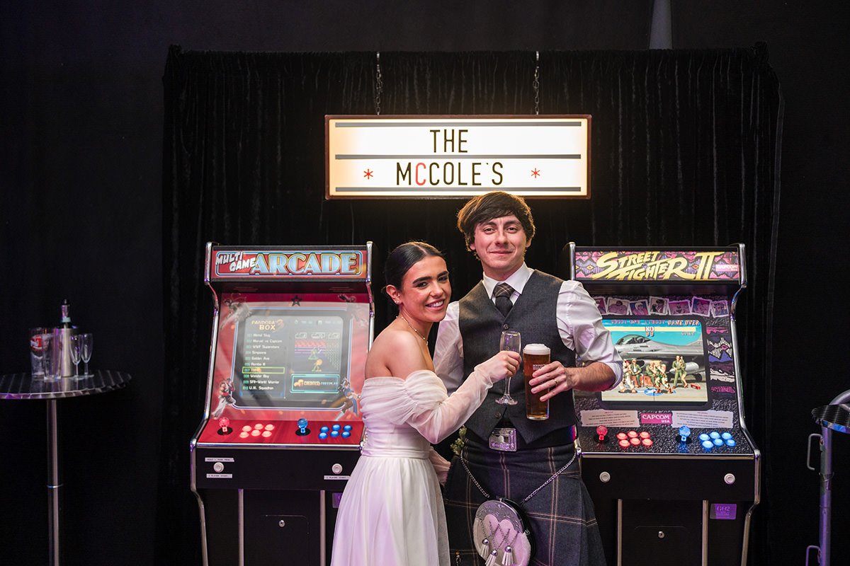 Bride and groom posing with drinks between retro arcade machines during their wedding reception in Glasgow