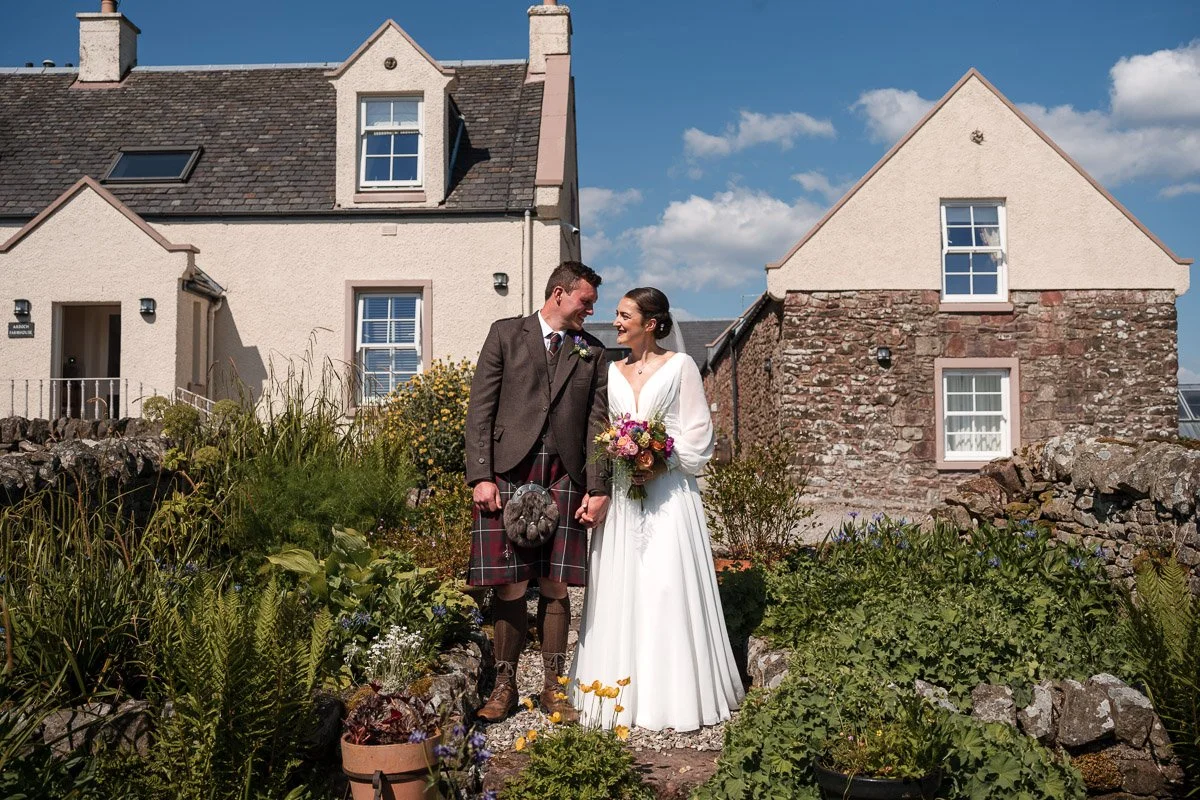 A bride and groom in wedding attire holding hands and smiling at each other outdoors in front of a house with a garden on a sunny day at the wedding at Ardoch Loch Lomond