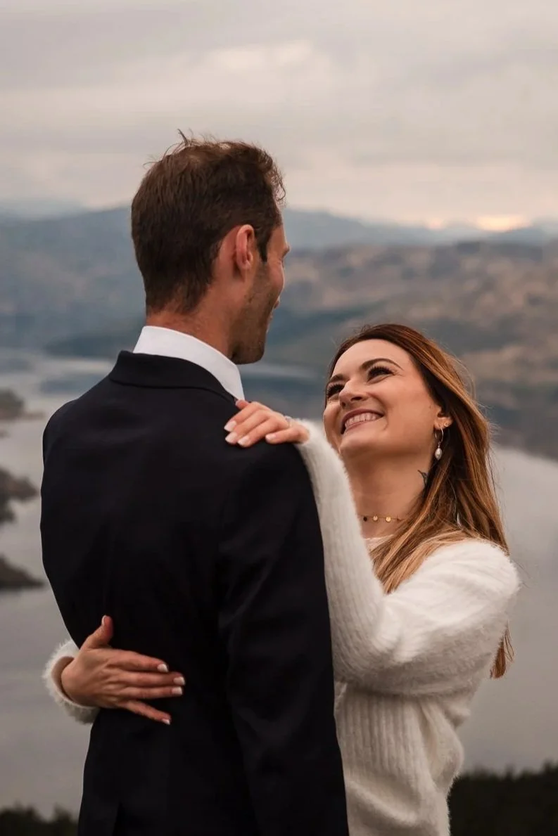 A couple smiling and embracing outdoors with mountains and a loch in the background atop Ben A'an with Loch Katrine in the background