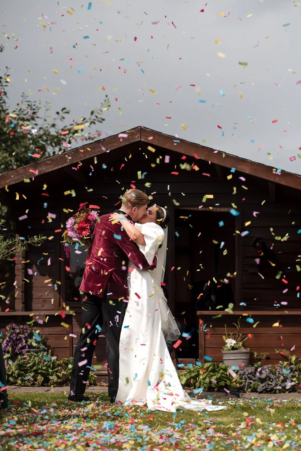A newlywed couple kissing under falling colorful confetti in front of a small wooden house surrounded by flowers and greenery.