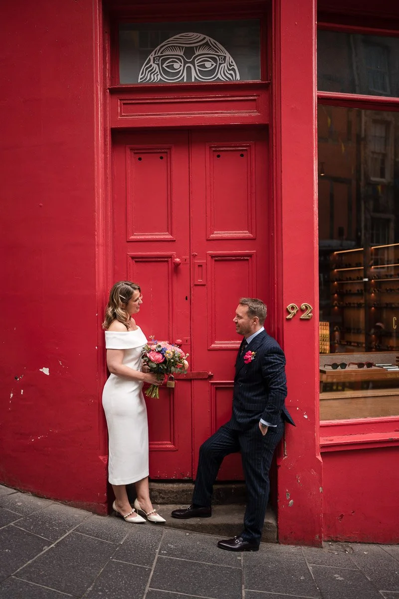 A couple dressed in wedding attire stands in front of a large red door with the number 92 on a building at an Edinburgh elopement. The woman is holding a bouquet of flowers, and the man is leaning against the wall, both smiling at each other.