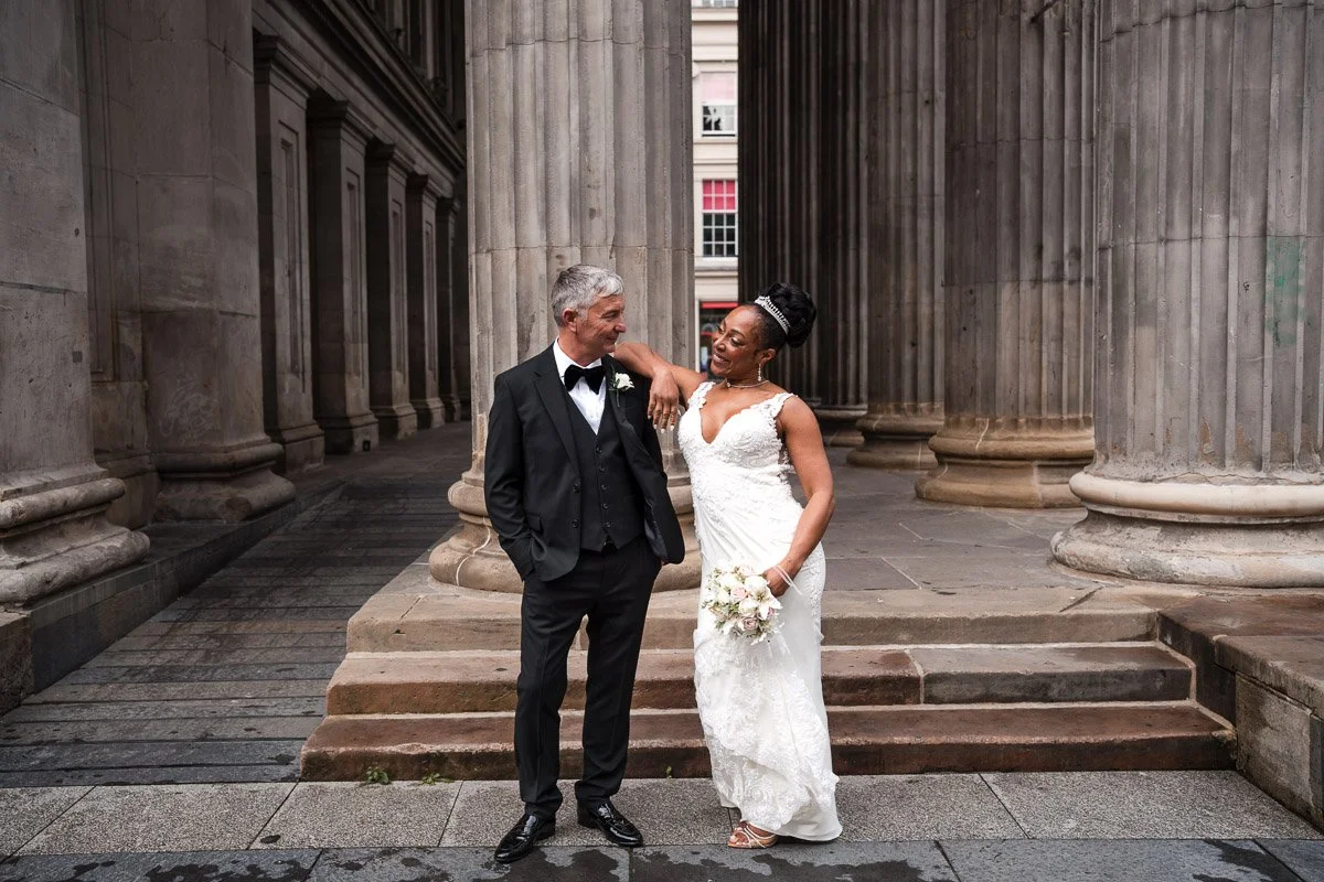 A bride and groom in wedding attire posing together outdoors in front of large stone columns of a historic building in Glasgow at their elopement