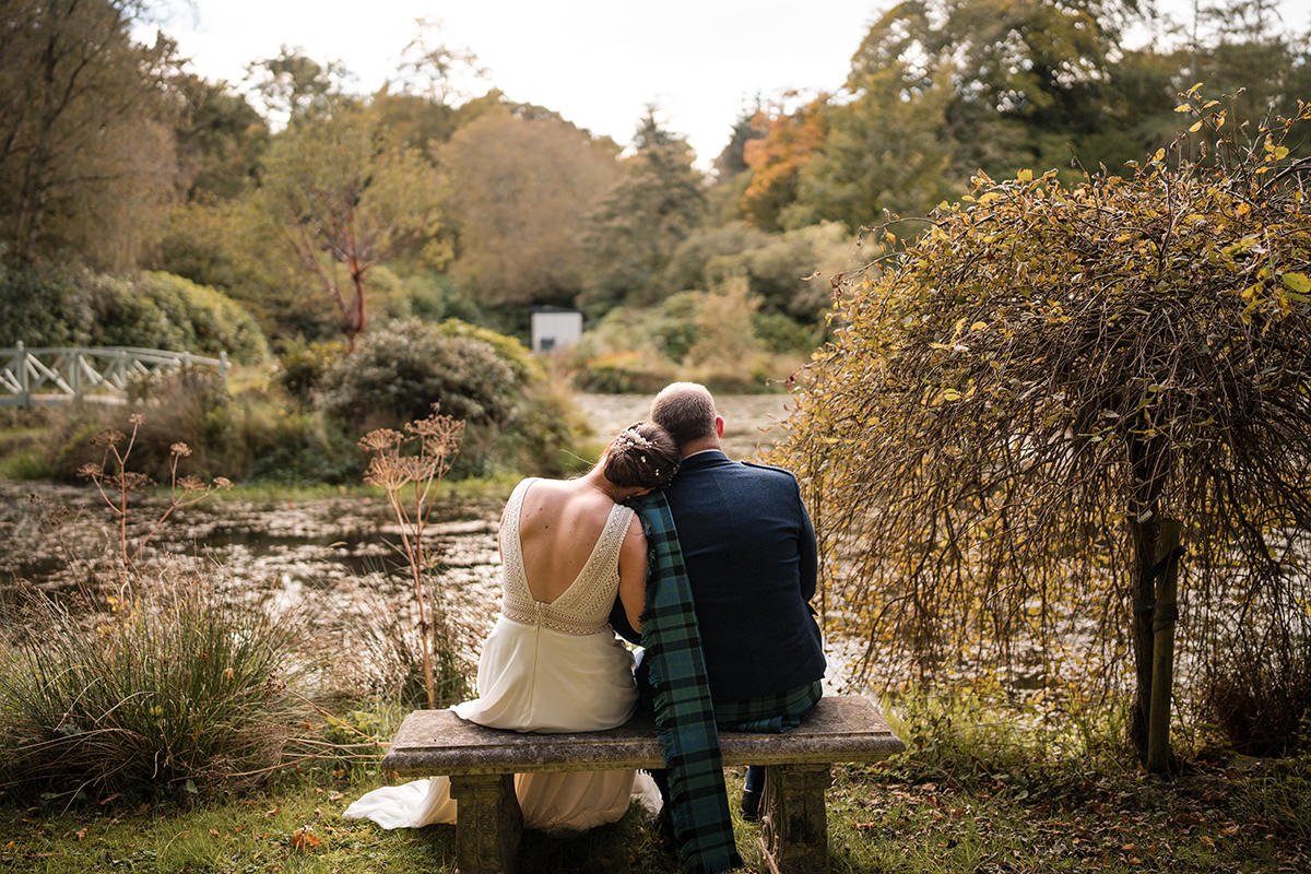A couple sitting on a park bench by a loch at Glencorse House, with trees and bushes in autumn foliage, embracing each other.