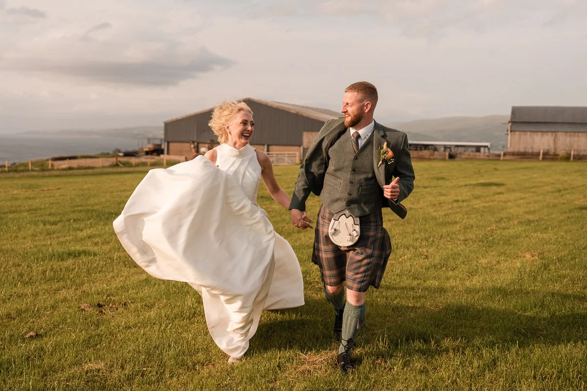 A bride and groom dressed in wedding attire running hand in hand across a grassy field with rustic buildings in the background whilst on Millport, Scotland