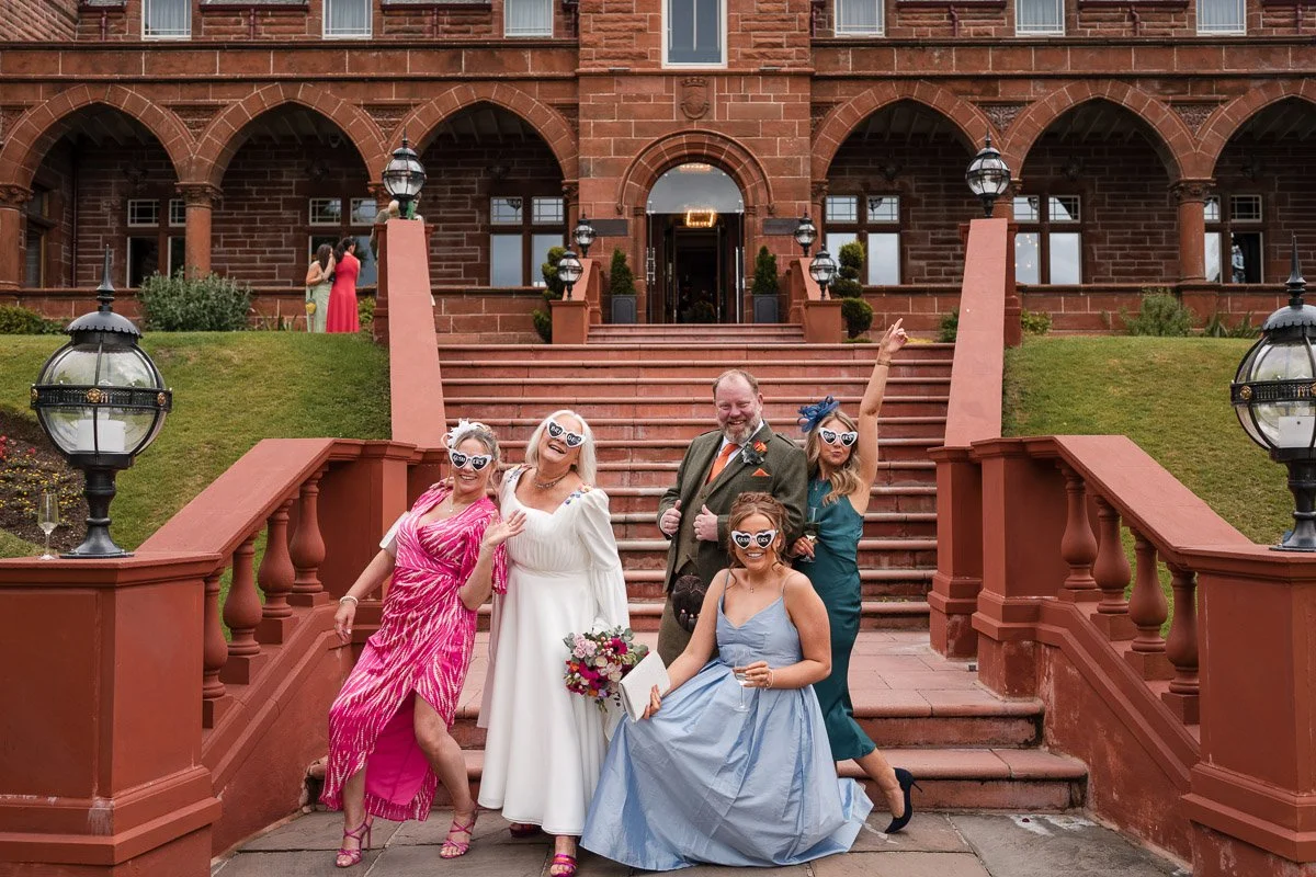 A group of five women and one man dressed in wedding attire, standing on the steps of a brick building with arched windows and red stone. The women are wearing colourful dresses and sunglasses, some holding drinks and a bouquet of flowers at Boclair
