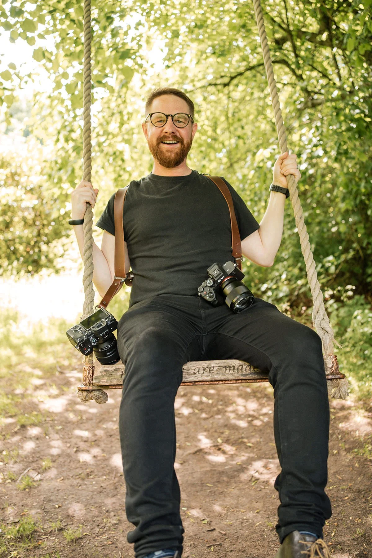 A man with glasses and a beard sitting on a wooden swing outdoors, holding the ropes, surrounded by green trees, with cameras hanging around his neck, smiling.