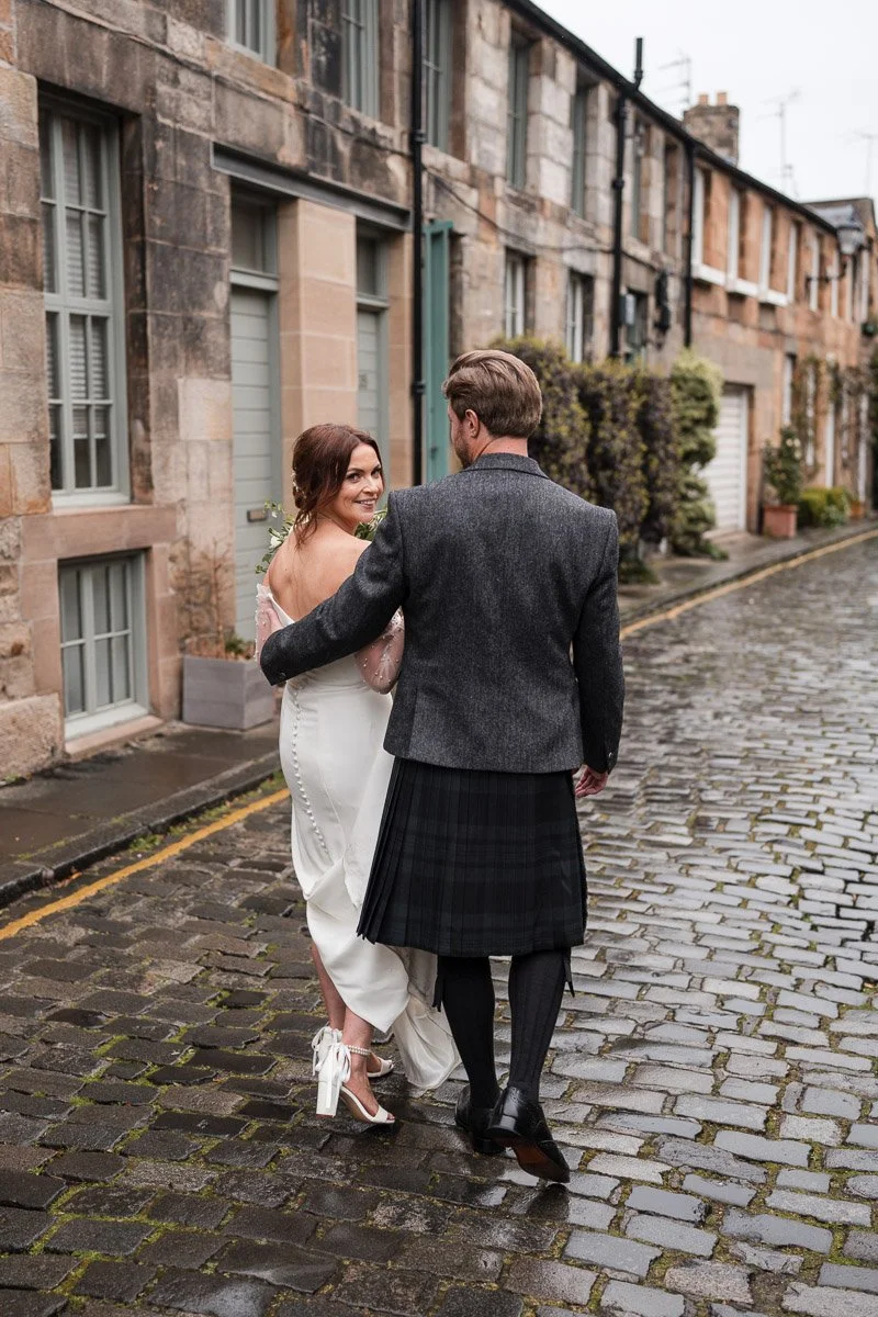 A couple walking on a cobblestone street, Circus Lane, with the woman in a wedding dress and the man in traditional Scottish attire, looking at each other and smiling after their Edinburgh city elopement