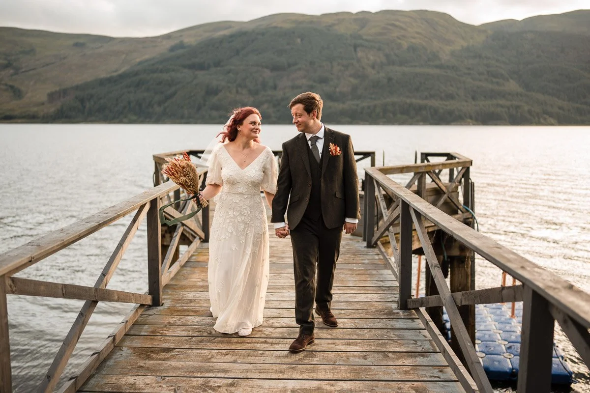 A bride and groom walking hand in hand on a wooden pier over a lake with green rolling hills in the background, during cloudy weather at the elopement in Dunoon
