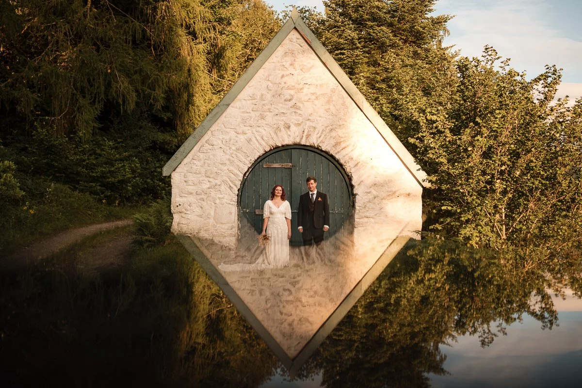 A bride and groom standing in front of a small white stone building with a rounded green door, reflected in a body of water at their elopement in Dunoon.