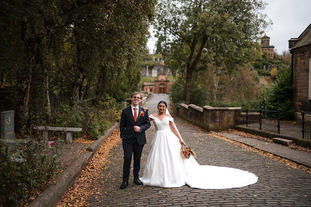 A bride and groom walking arm in arm on a cobblestone path, surrounded by trees and autumn leaves, with historic buildings in the background at a Glasgow city elopement