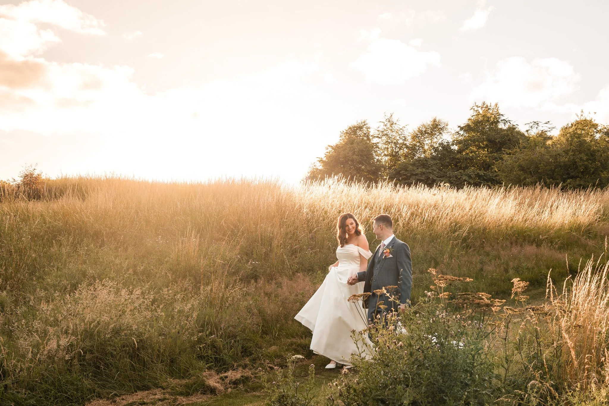 Wedding couple walking hand in hand through a grassy field at sunset, with trees in the background.