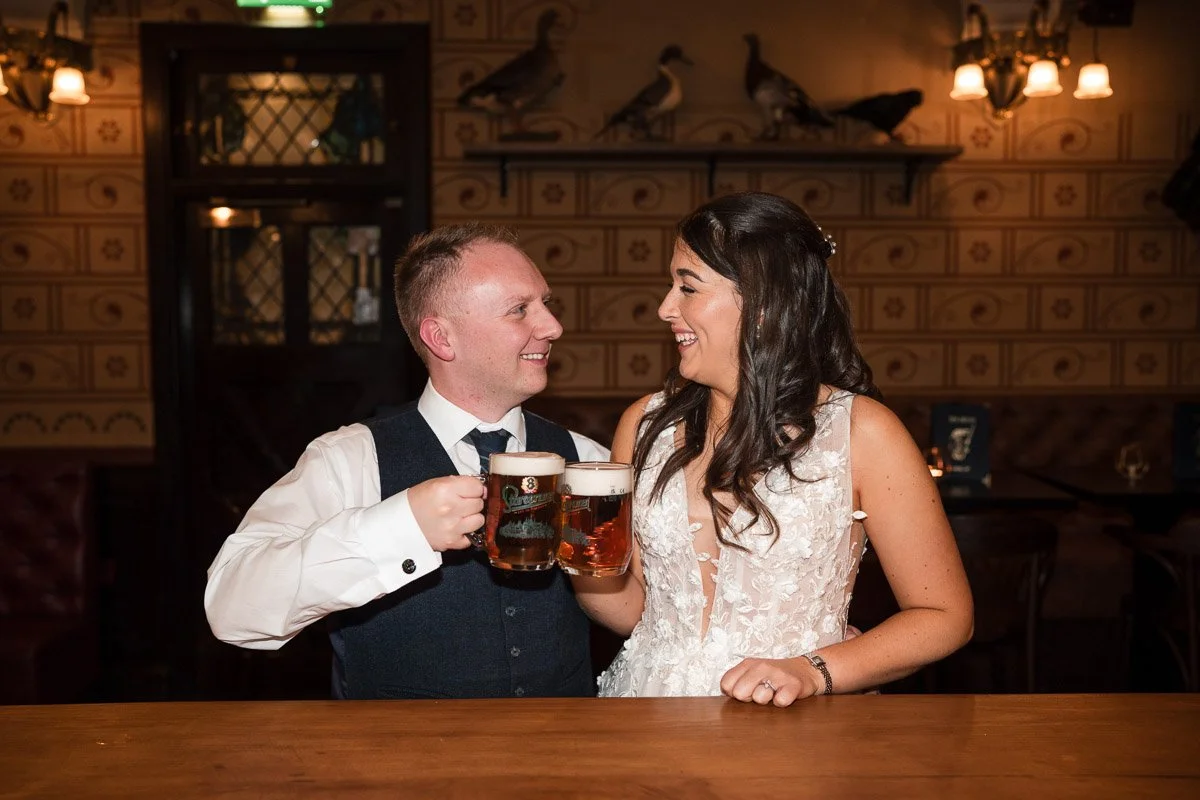 A bride and groom in wedding attire smiling at each other while holding beer mugs in a bar or pub setting with wooden decor and dim lighting at their wedding at The Kelvin in Glasgow
