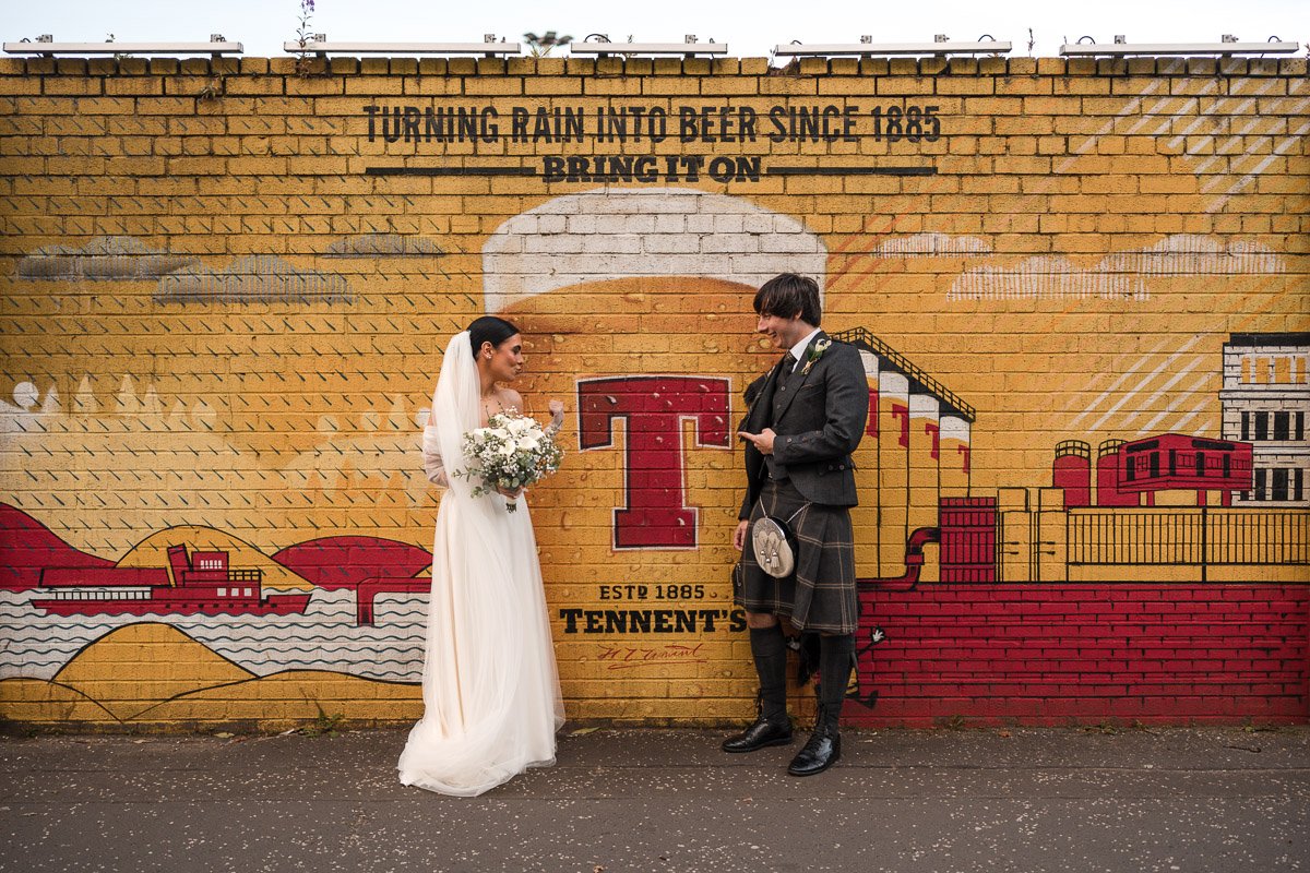 Bride in a white wedding dress holding a bouquet of flowers and groom in a kilt with a sporran, standing in front of a colourful Tennents mural on a brick wall in Glasgow.