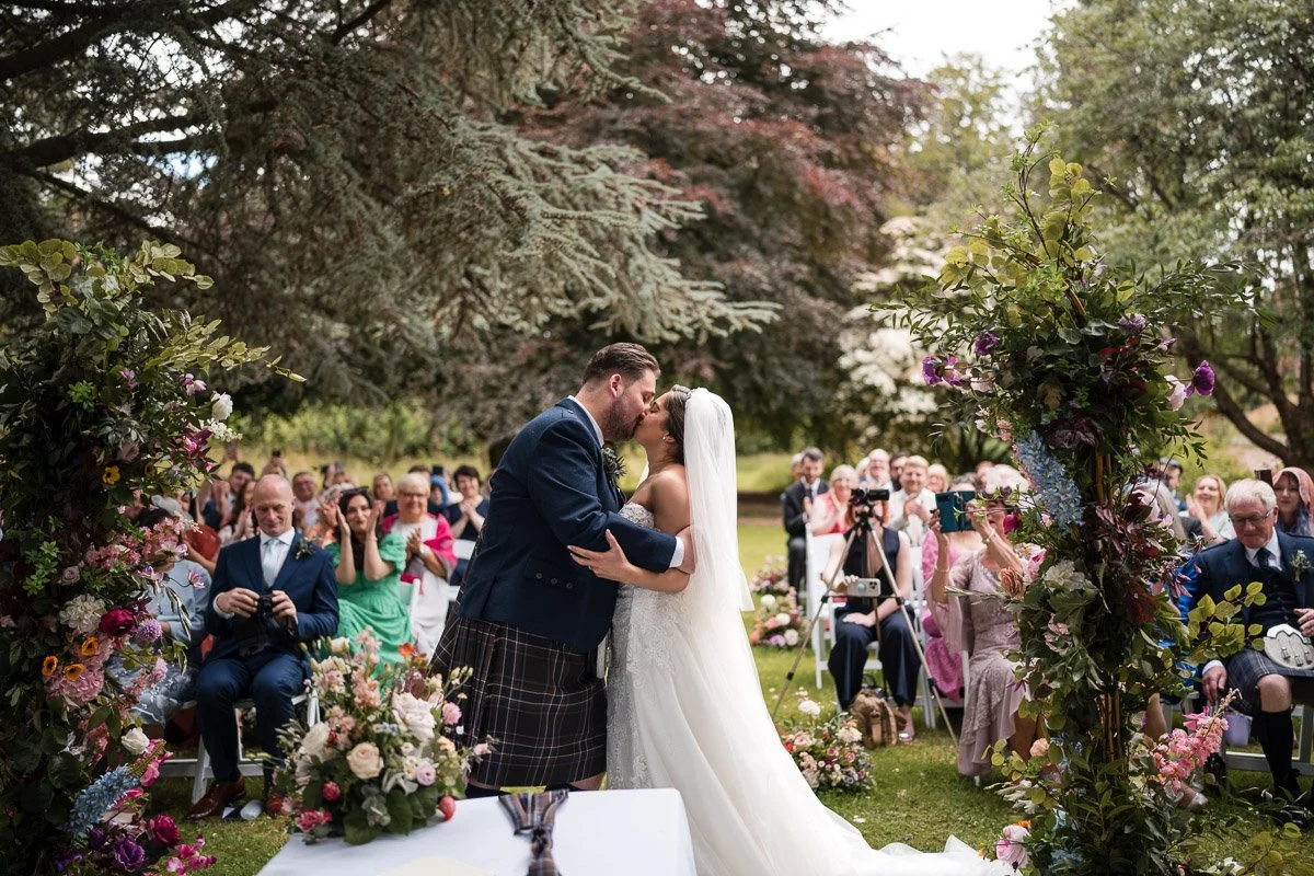 A wedding ceremony outdoors with a bride and groom kissing under a floral arch, surrounded by seated guests on a lush green lawn with large trees in the background at an outdoor ceremony at Rufflets, St. Andrews
