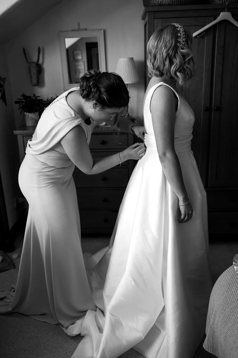 A bride getting help adjusting her wedding dress while a woman assists her in a bedroom.