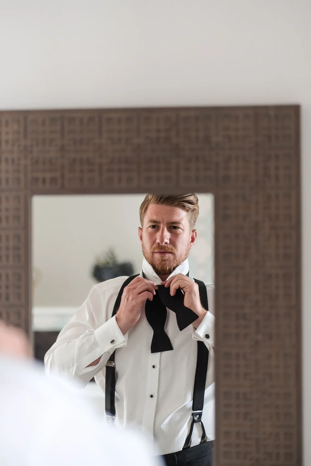 A man is looking at himself in a mirror, adjusting his black bow tie. He is dressed in a white dress shirt and black suspenders.