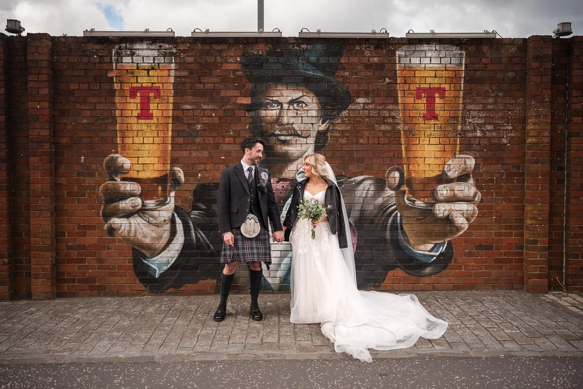 A newlywed couple after their Glasgow elopement standing in front of a Tennents mural of a man holding two glasses with the letter T on each glass. The man in the mural is wearing a top hat and suit, and is smiling.