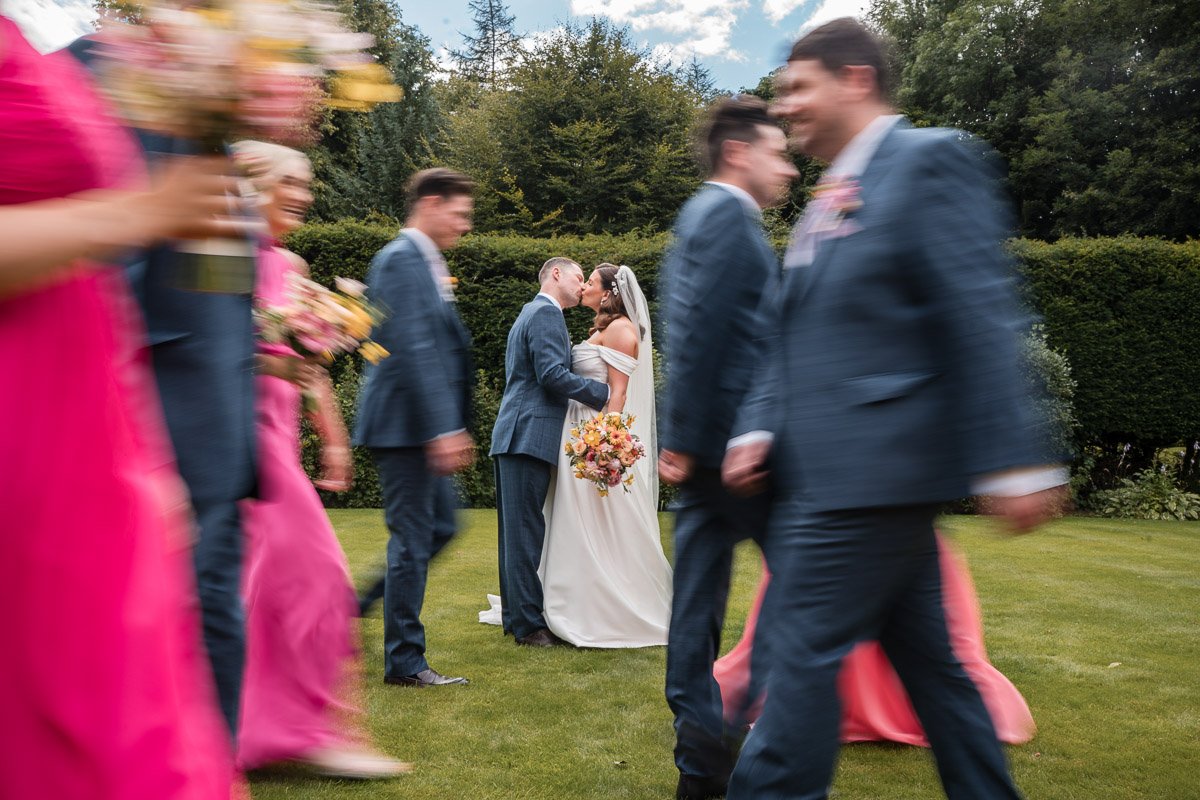 Bride and groom kissing in the background while wedding guests walk by in front, some holding bouquets at Balbirnie
