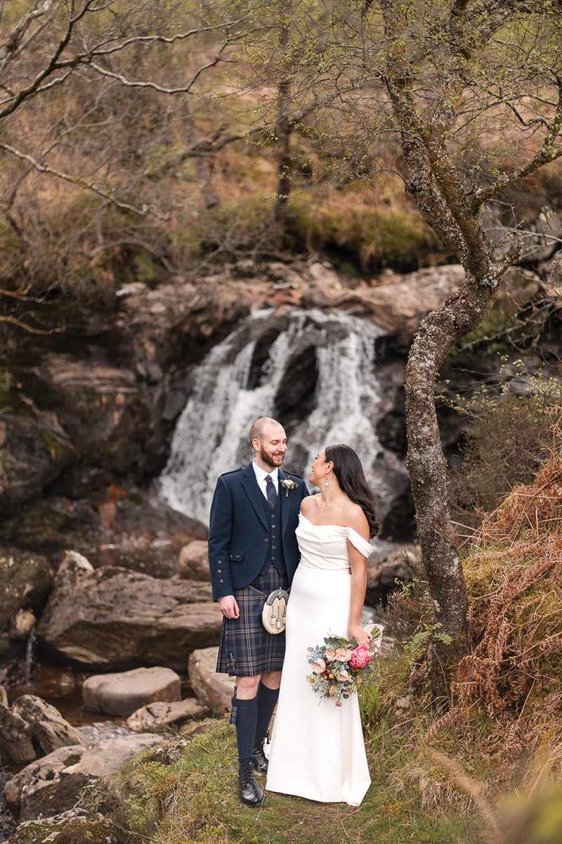 A bride and groom stand by a stream with a small waterfall in a wooded area during autumn at Carrick Castle. The bride is in a white off-shoulder wedding dress holding a bouquet, and the groom is in traditional Scottish attire including a kilt.