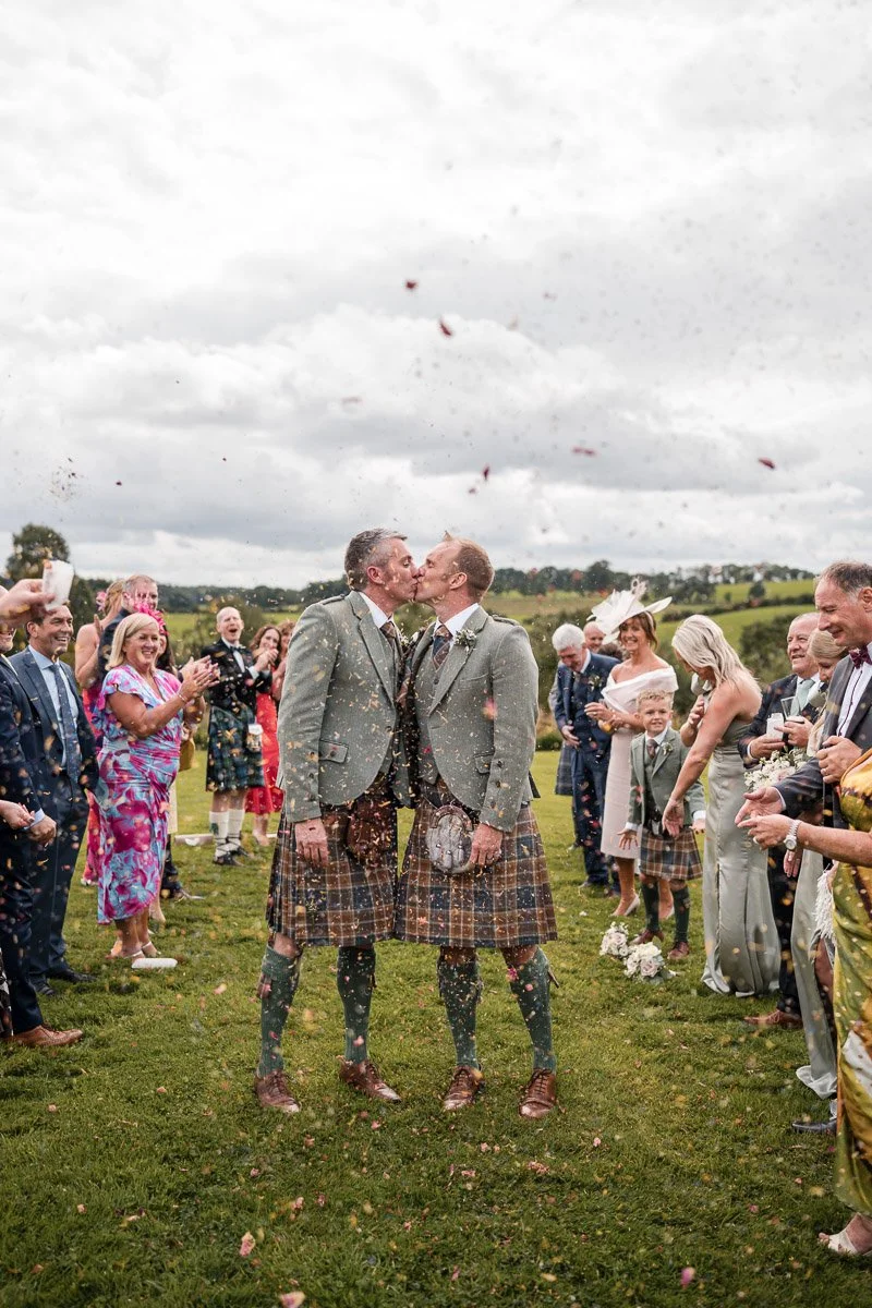 Two grooms dressed in traditional Scottish kilts kissing at a wedding ceremony; guests surrounding them cheering and clapping outdoors on a cloudy day.