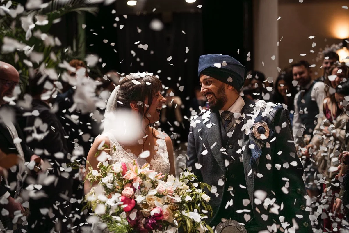 A bride and groom celebrating their wedding with guests, confetti in the air, the bride holding a large bouquet of flowers, both smiling and surrounded by friends and family whilst at their wedding at Engine Works, Glasgow