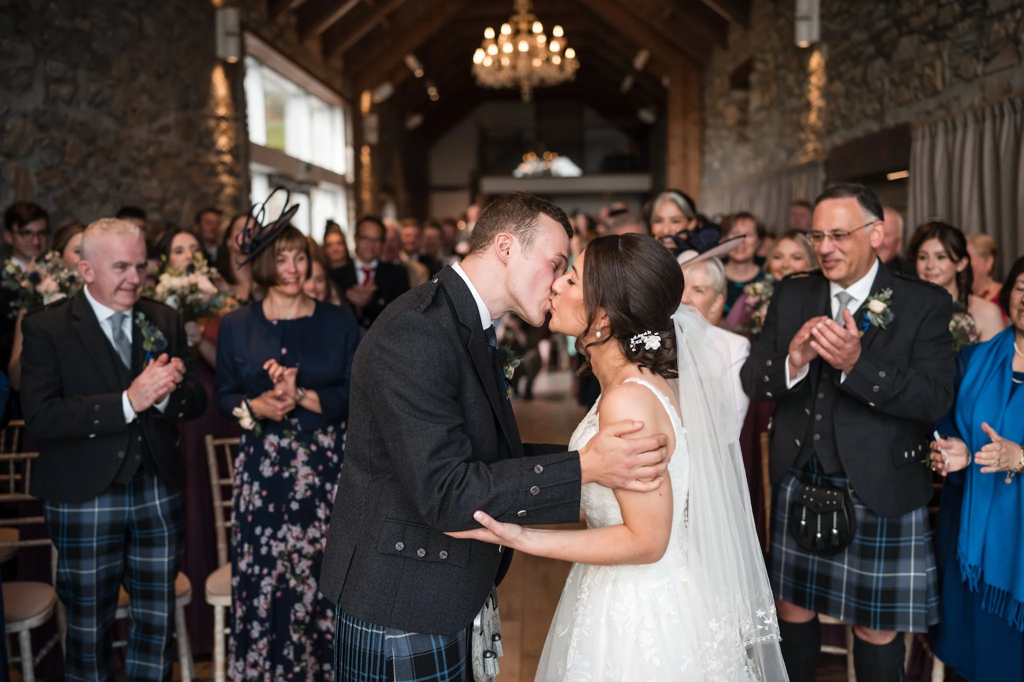 A wedding ceremony with a bride and groom kissing, surrounded by family and friends in a rustic indoor venue with wooden beams, stone walls, and a chandelier at GG's Yard