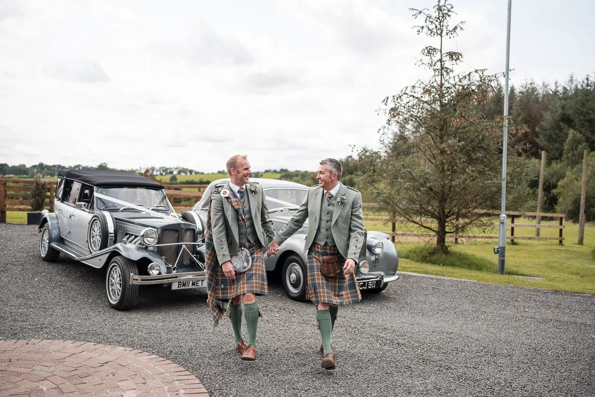 Two grooms dressed in traditional Scottish attire, including kilts, walking hand-in-hand outdoors near vintage cars, smiling at each other on a cloudy day at Eden Leisure Village