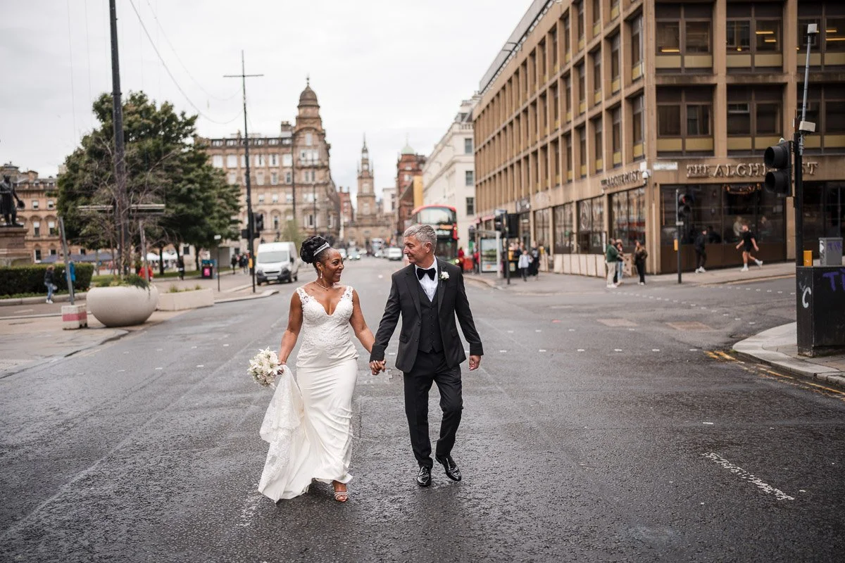 A newlywed couple walking hand in hand on a city street, with buildings, pedestrians, and a double-decker bus in the background in a Glasgow city elopement